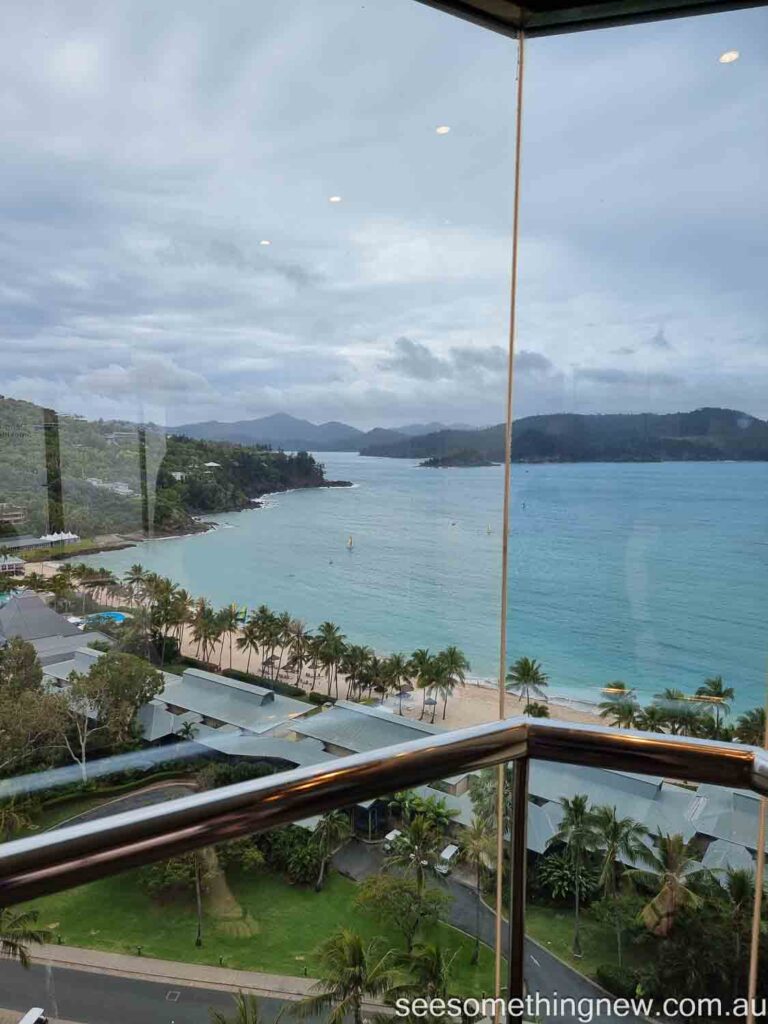 View from the Hamilton Island Reef View Hotel Lift of palm tree fringed beach with an azure sea behind it and mountan islands in the background.