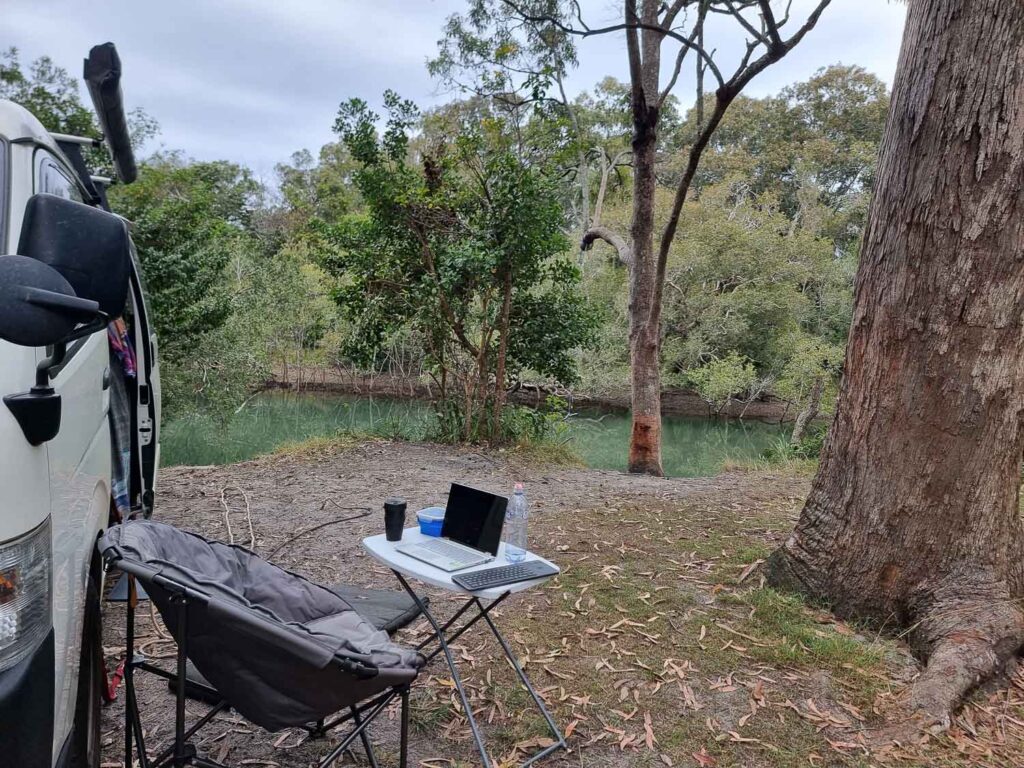 Folding Chair and table with laptop with side view of van looking over sugar mill creek which has bushes and aqua green coloured water