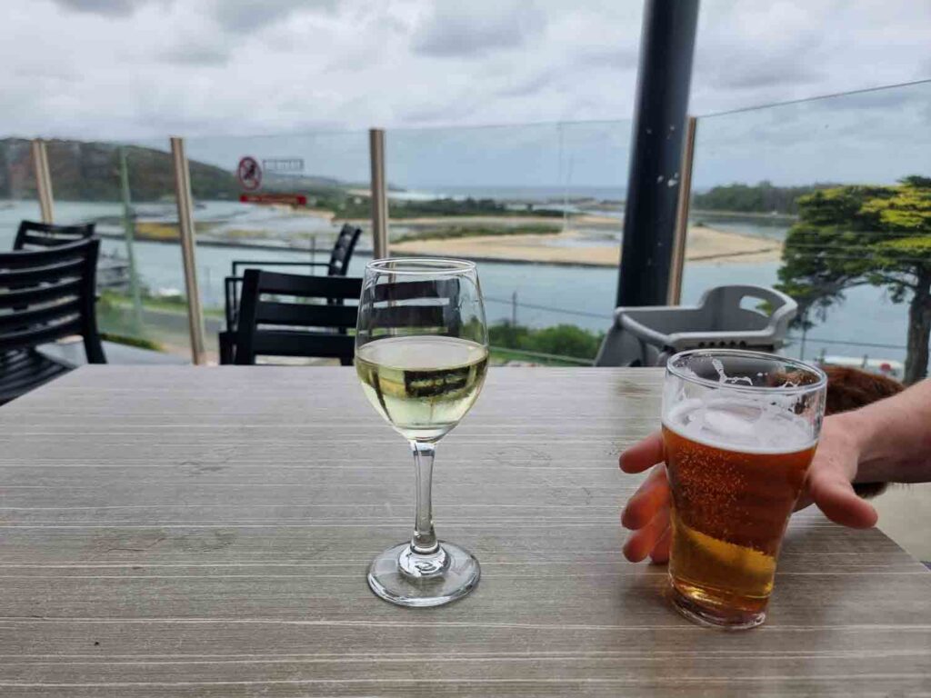 A wine and beer on a table with a view of an inlet through class barricade of deck
